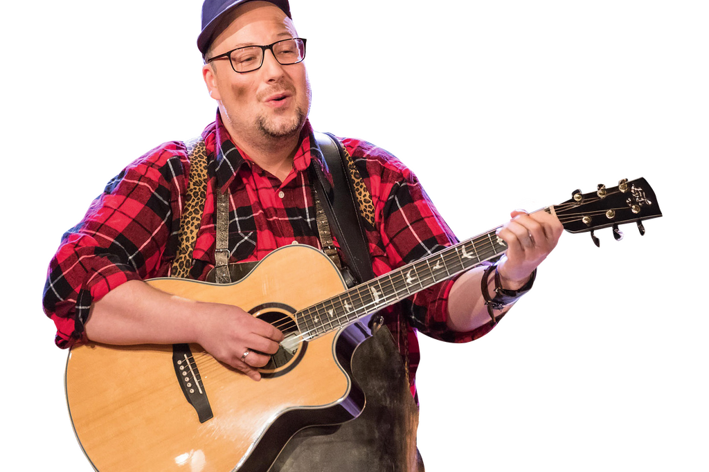 Ein Musiker mit Gitarre singt fröhlich in einem karierten Hemd. | © DerSchmied