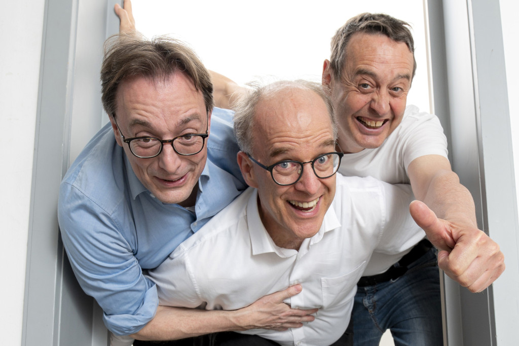 Drei Männer posieren lachend zusammen in einem hellen Raum. | © Nico Manger
