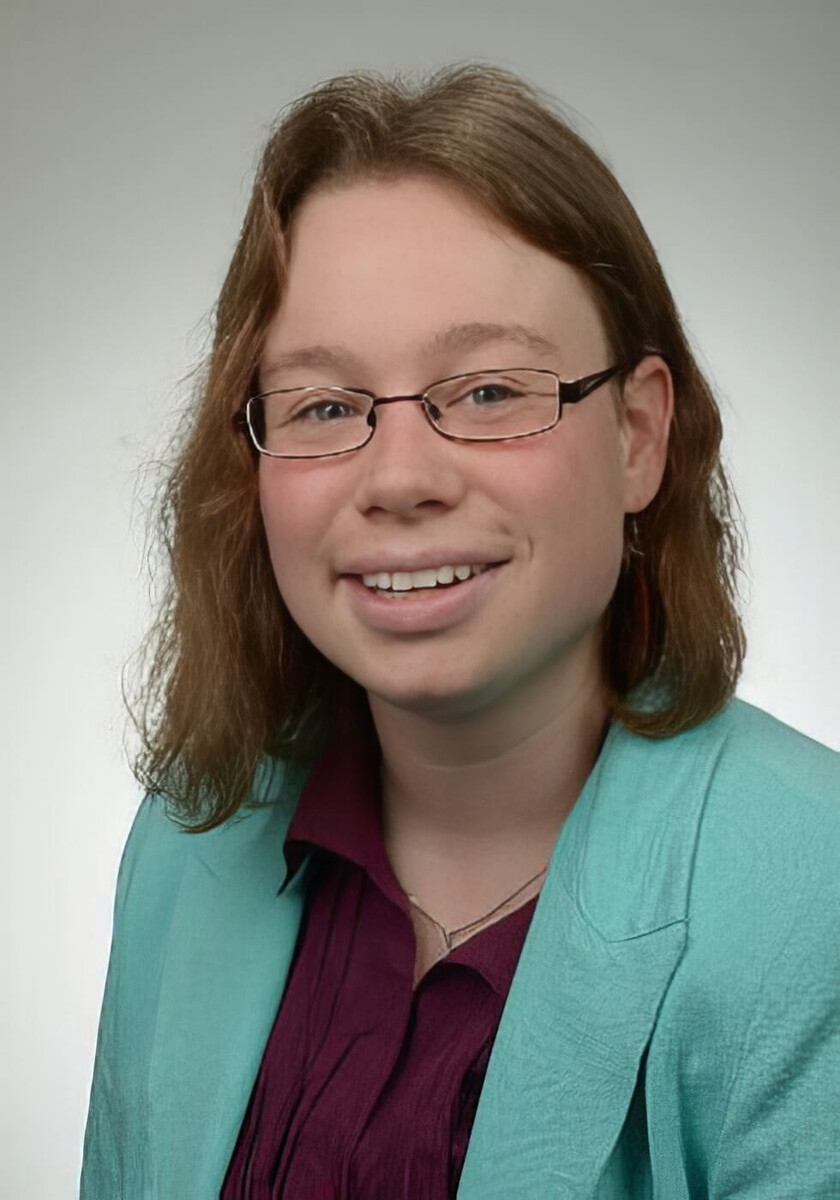Julya Berzen lächelt freundlich in einem hellen Blazer und sitzt vor neutralem Hintergrund.